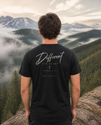 Fictional model wearing the Different Romans 12:2 Tee – Back Print in Black, back view, Pacific Northwest scene (digitally composited lifestyle image)