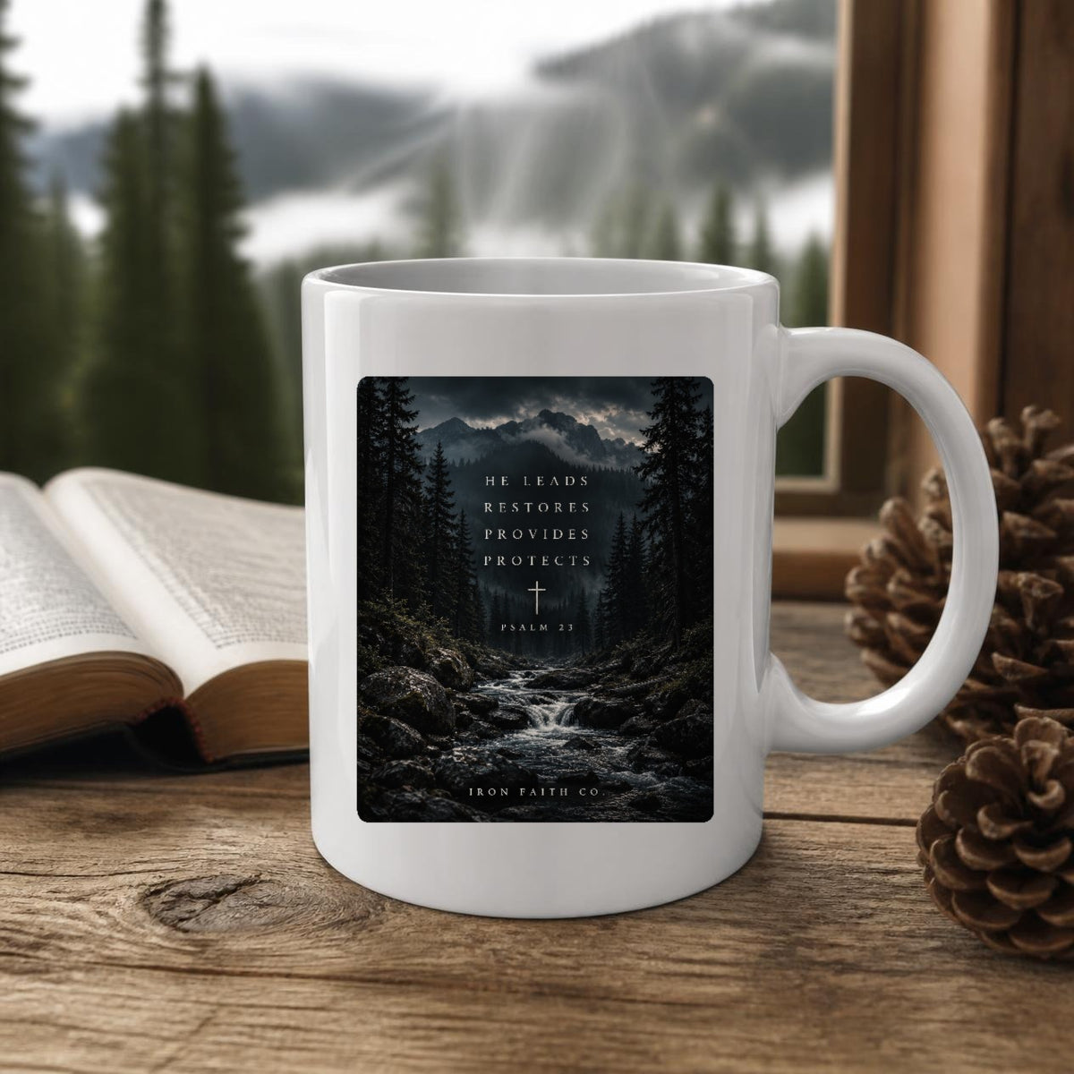 The He Leads Psalm 23 Mug (11oz, white) on a wooden cabin table with an open Bible and pine cones (digitally composited scene image)