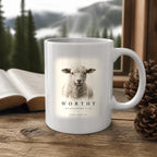 The Worthy Revelation 5:12 Mug (11oz, white) on a wooden cabin table with an open Bible and pine cones (digitally composited scene image)