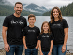 Lifestyle image (digitally composited, real print artwork at true print size): Christ Is King tees on a fictional family of four at a Pacific Northwest lake (adult, women's and youth sizes)
