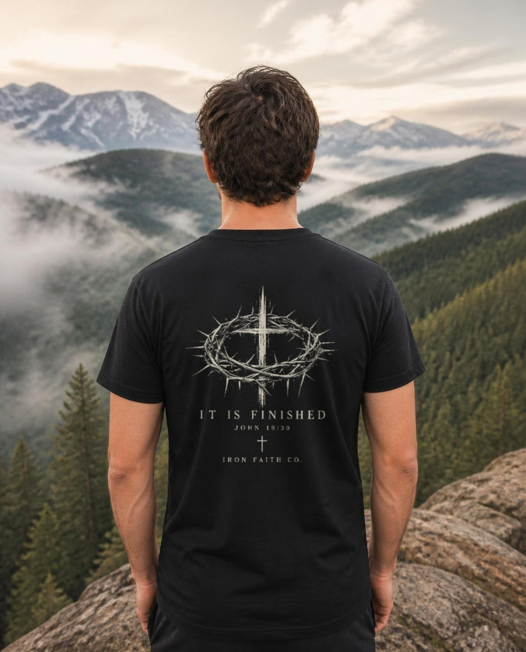 Lifestyle image (digitally composited, real print artwork at true print size): Back of the It Is Finished tee (full design on back) on a fictional model overlooking misty mountains