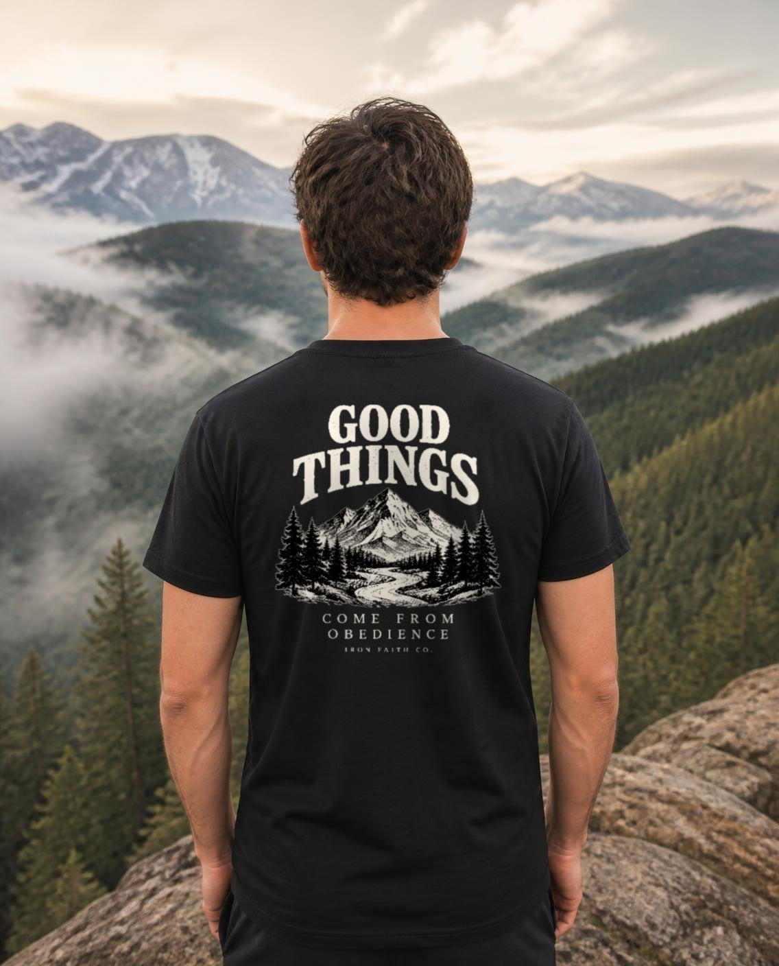 Fictional model wearing the Good Things Come From Obedience Tee – Back Print in Black, back view, Pacific Northwest scene (digitally composited lifestyle image)