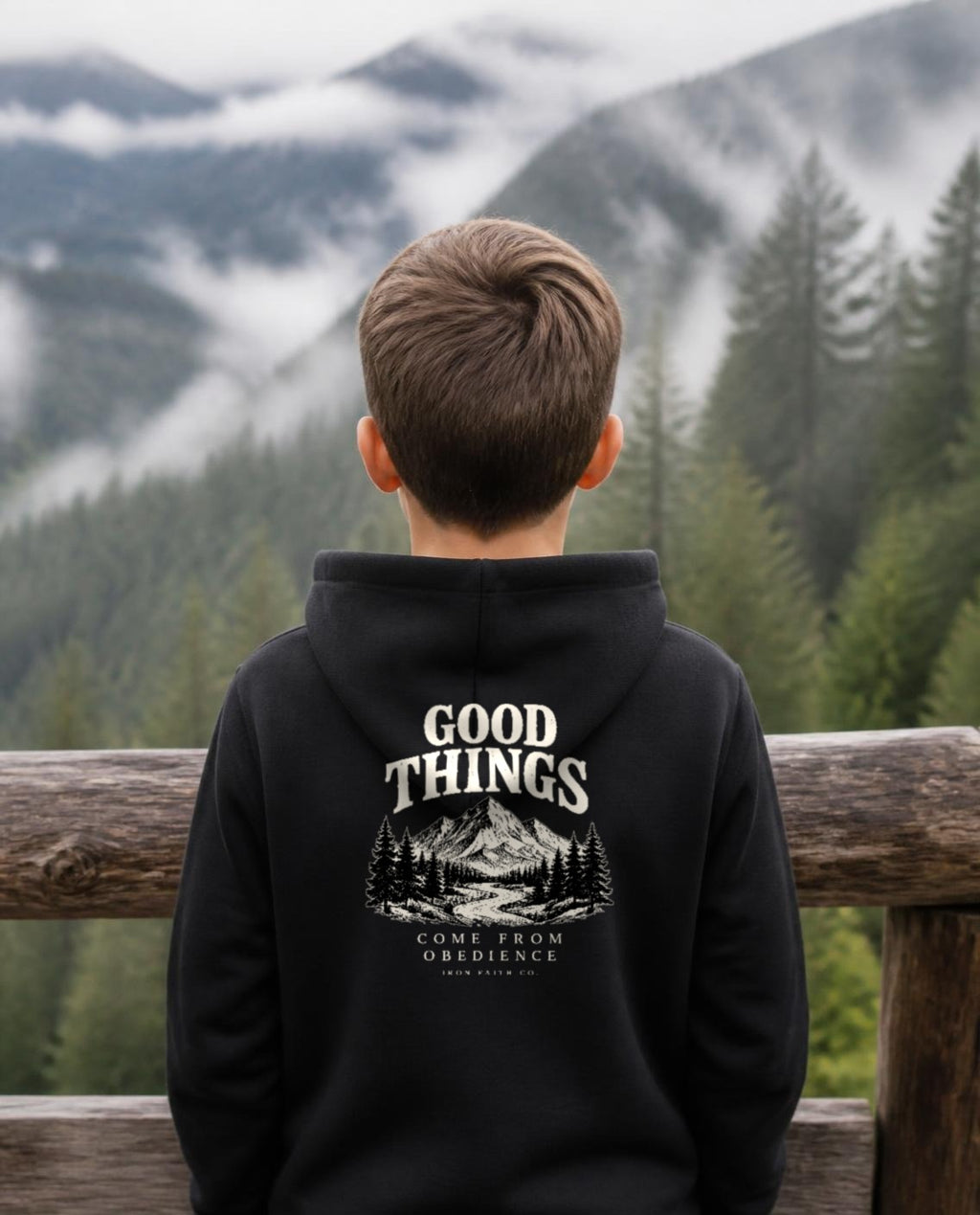 Fictional model wearing the Good Things Come From Obedience Youth Hoodie – Back Print in Black, back view, Pacific Northwest scene (digitally composited lifestyle image)
