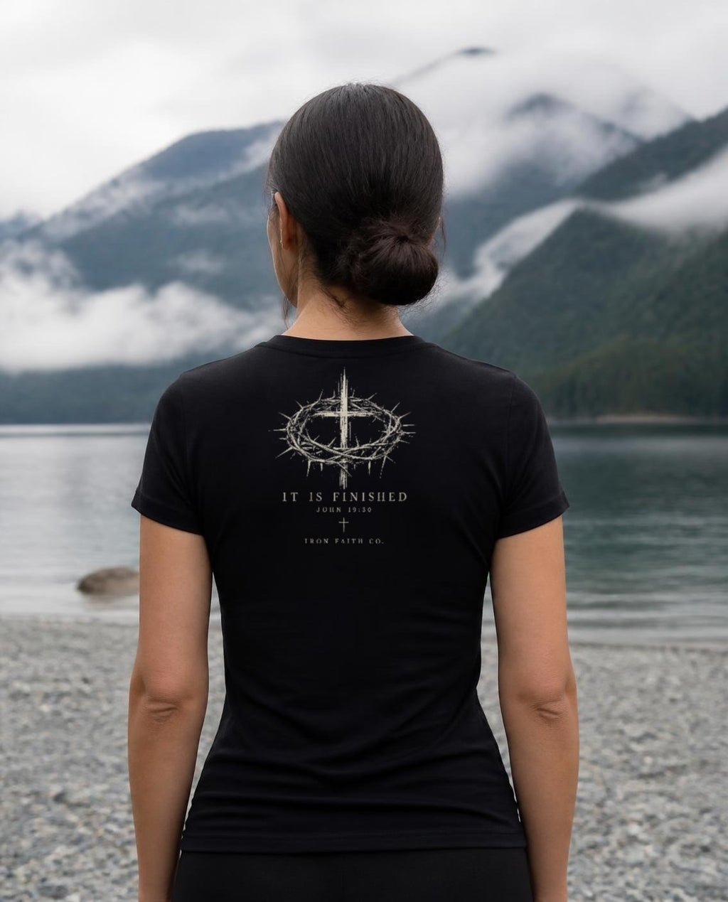 Fictional model wearing the It Is Finished Women's Fitted Tee in Black, back view, Pacific Northwest scene (digitally composited lifestyle image)