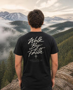 Fictional model wearing the Walk By Faith Tee – Back Print in Black, back view, Pacific Northwest scene (digitally composited lifestyle image)