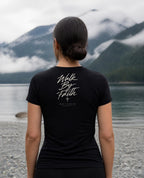 Fictional model wearing the Walk By Faith Women's Fitted Tee – Back Print in Black, back view, Pacific Northwest scene (digitally composited lifestyle image)