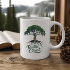 Rooted in Christ Mug (white ceramic) on a wooden table by a window with a Bible and pinecones – digital mockup